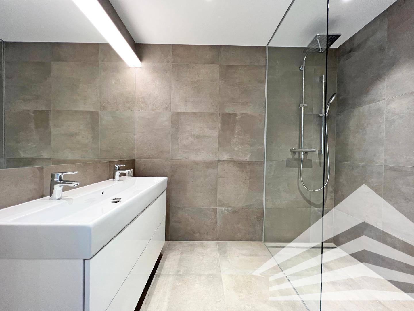 Modernes Badezimmer mit Doppelwaschbecken, großen grauen Fliesen und einer begehbaren Dusche.
