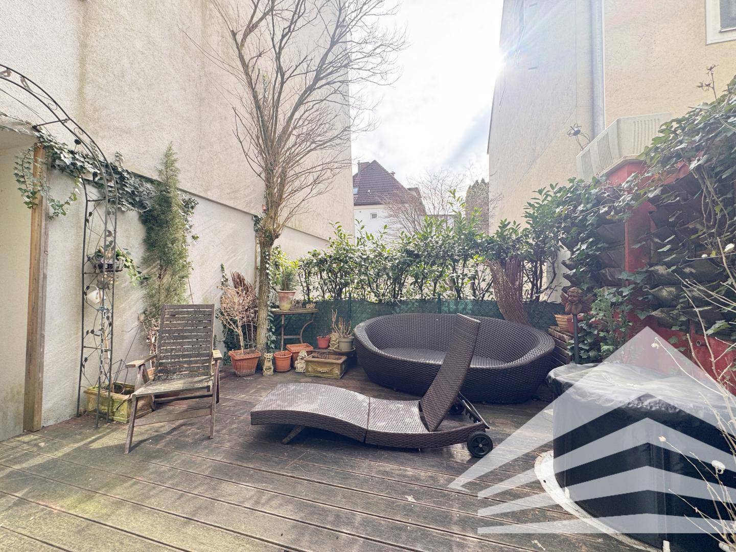 Sonnige Außenterrasse mit Holzdeck, Lounge-Möbeln und üppiger Bepflanzung.