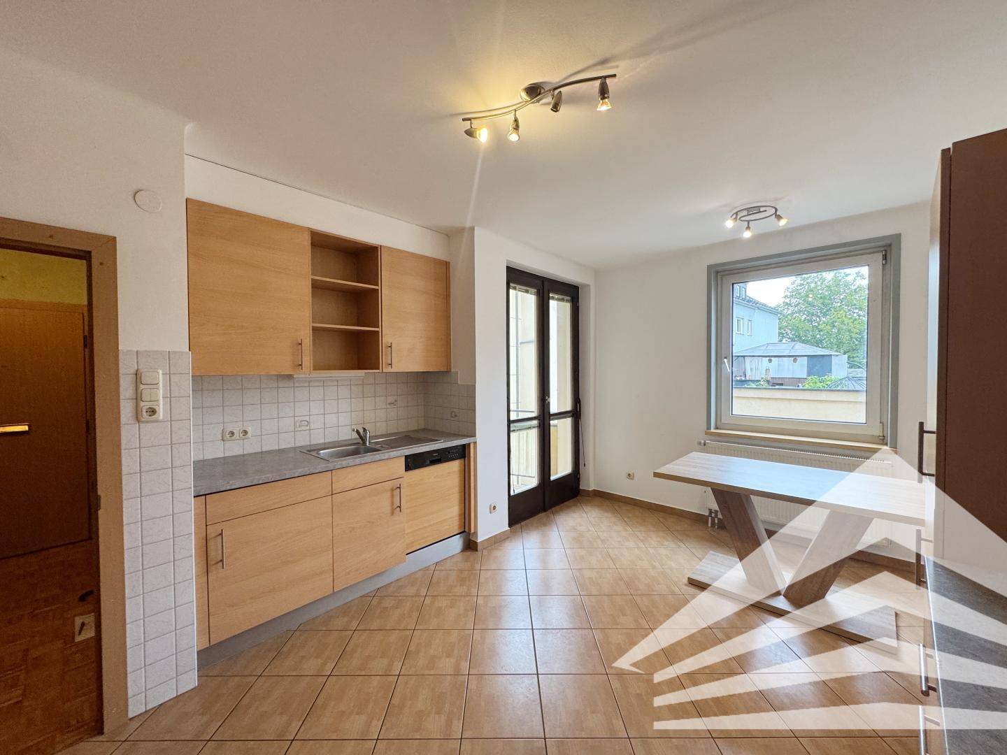Helle Küche mit Holzschränken, Fliesenboden, Spüle, Geschirrspüler und Zugang zum Balkon.