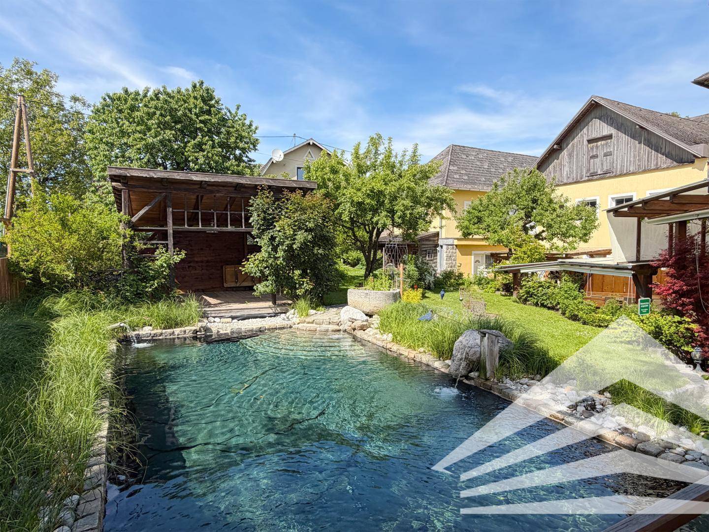 Natürlicher Schwimmteich mit klarem Wasser, umgeben von Grünflächen und einem rustikalen Holzhaus.