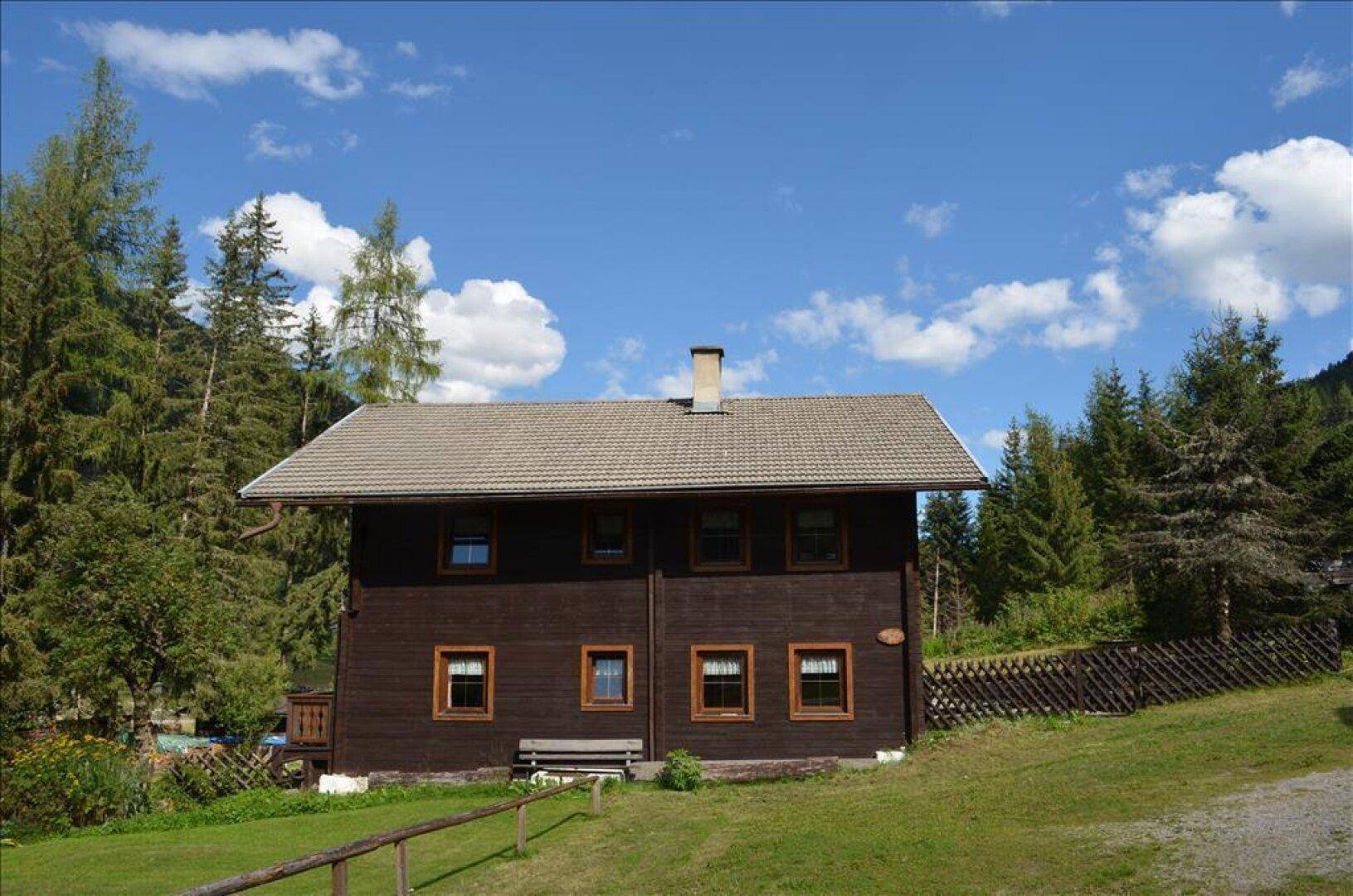 Rustikales Holzhaus mit Satteldach, umgeben von grünen Wiesen und dichtem Wald unter blauem Himmel.