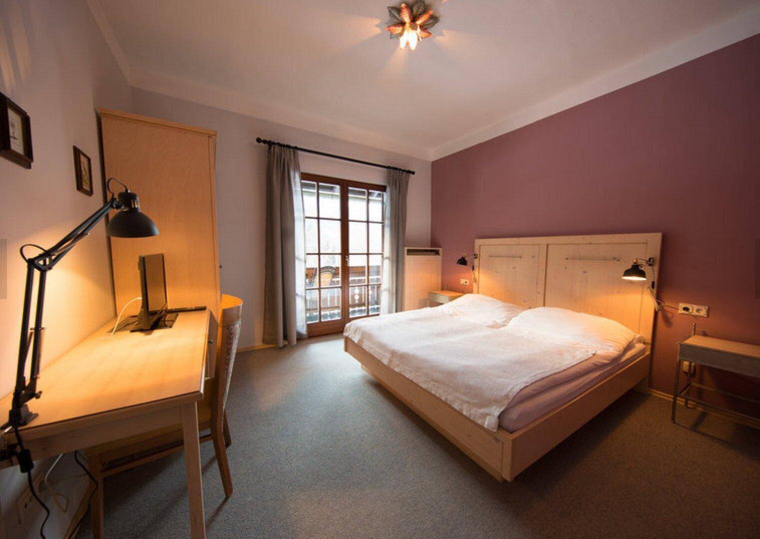 Schlafzimmer mit Doppelbett, Schreibtisch und Zugang zu einem Balkon, beleuchtet von natürlichem Licht.