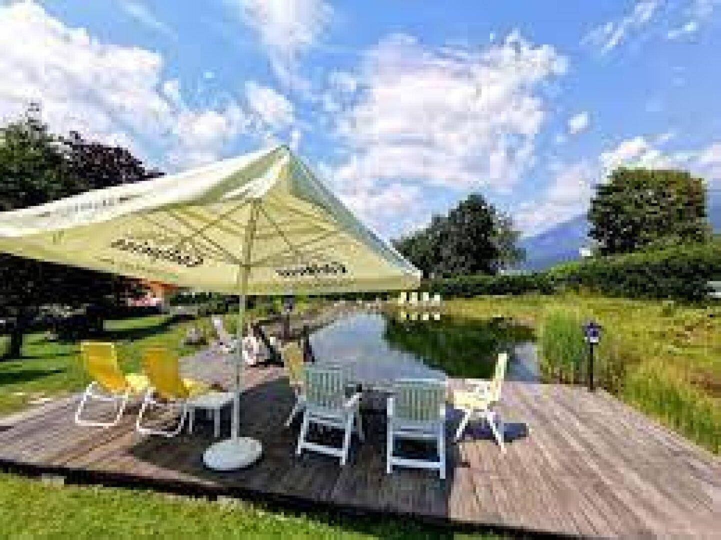 Entspannungsbereich im Freien mit Sonnenschirm, Sitzgelegenheiten und einem natürlichen Teich im Hintergrund.