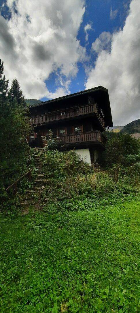 Alpines Holzhaus mit mehreren Balkonen, eingebettet in eine grüne Hügellandschaft unter dramatischem Himmel.