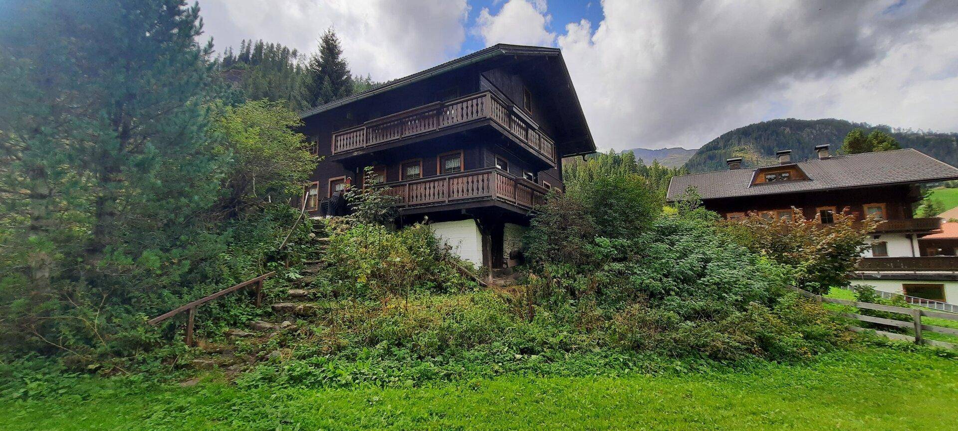 Traditionelles Holzchalet in alpiner Umgebung mit angrenzendem Nachbarhaus und üppiger Vegetation.