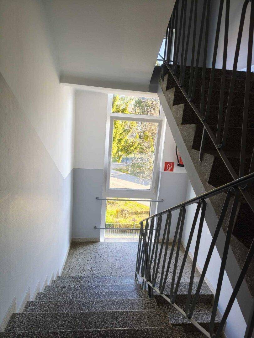 Treppenhaus mit Granitstufen und großem Fenster, das viel Tageslicht hereinlässt.