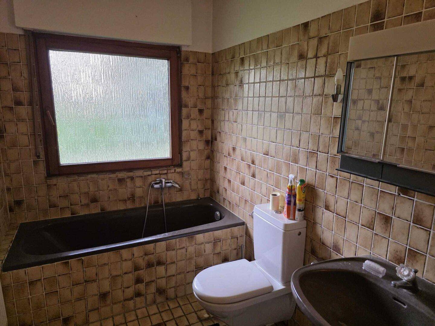 Badezimmer mit braunen Fliesen, Badewanne, Toilette und Fenster.