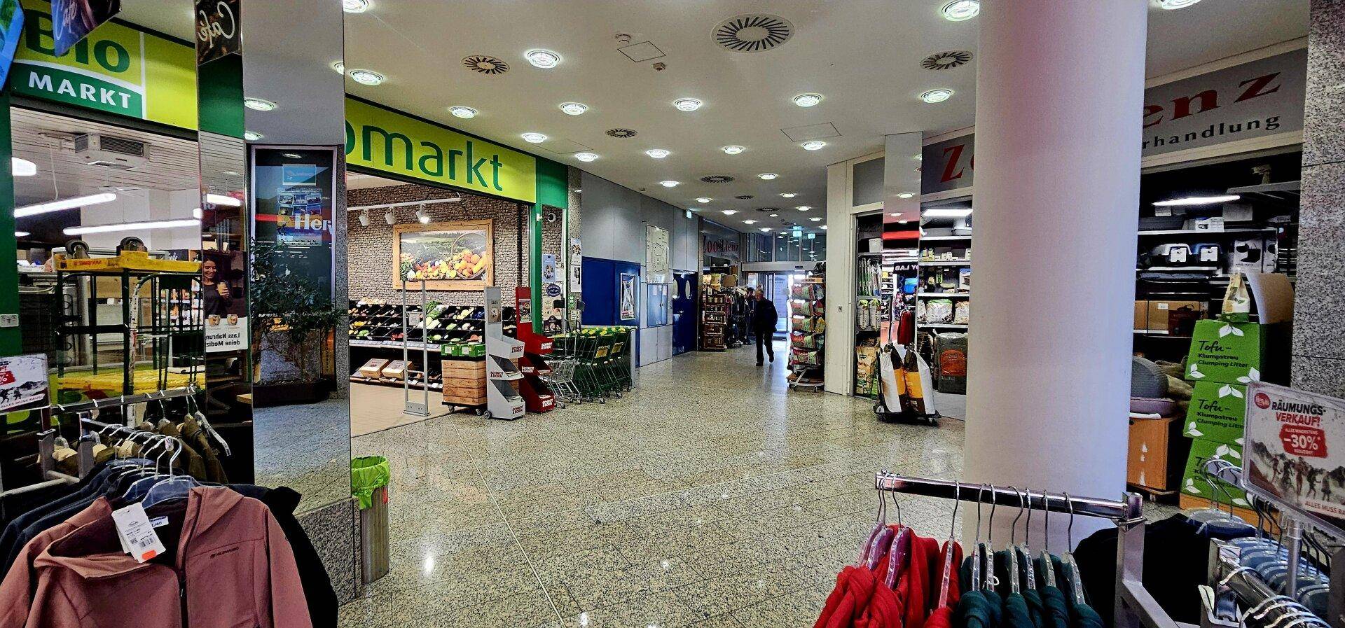 Innenansicht eines Einkaufszentrums mit verschiedenen Ladenfronten und Kleiderständern im Vordergrund.