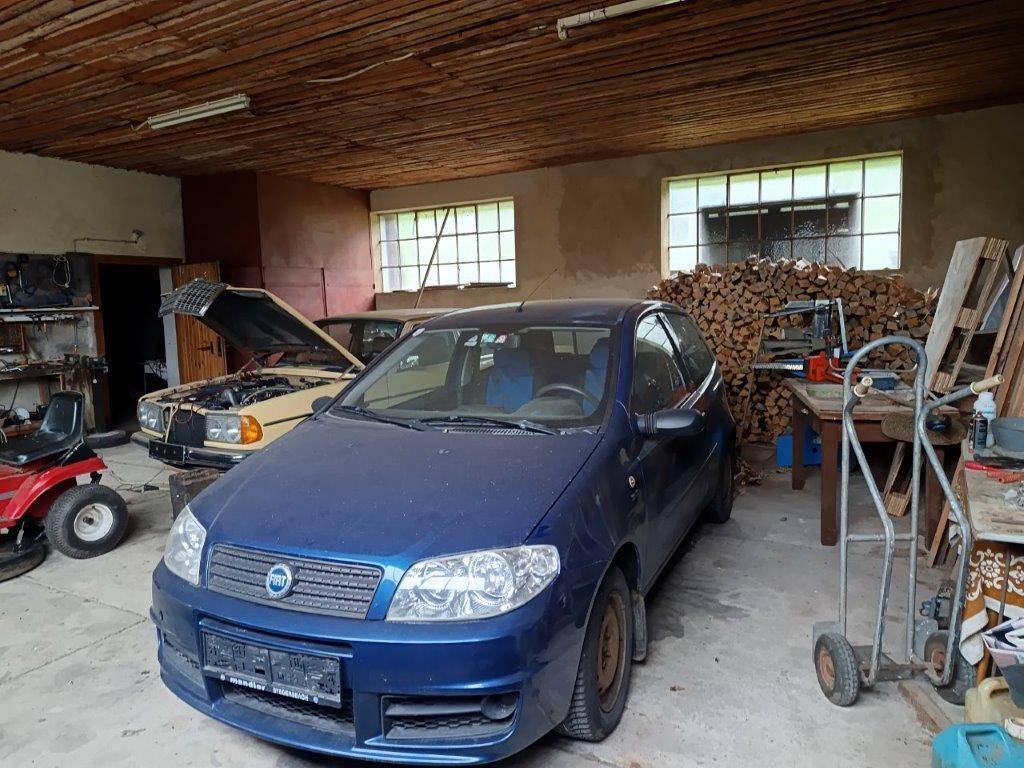 Innenansicht einer Garage mit zwei Autos, Werkzeugen und einem Stapel Brennholz unter einer Holzplankendecke.