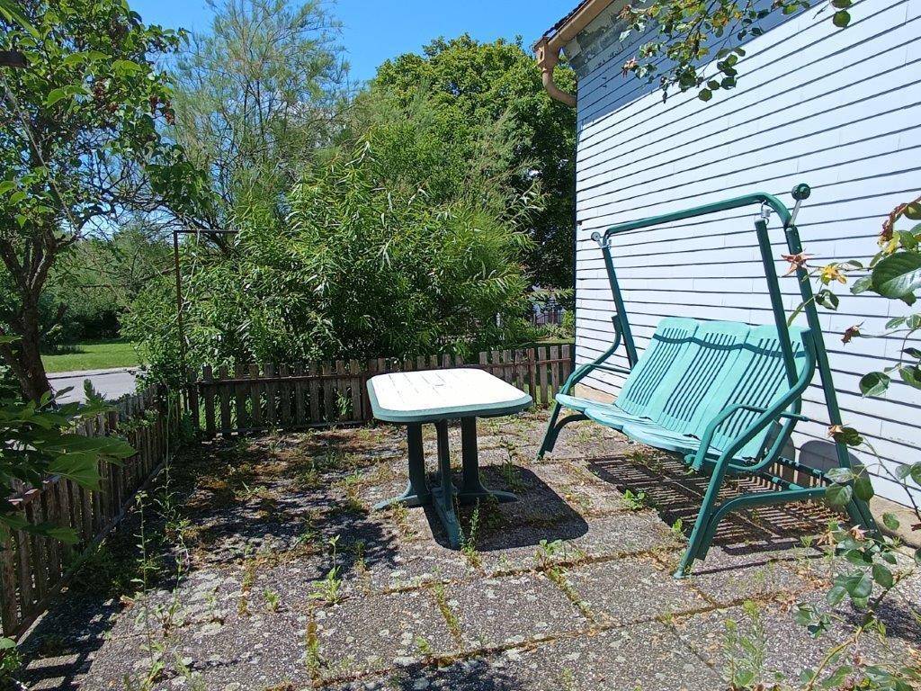 Kleine gepflasterte Terrasse mit Gartenschaukel und Tisch, umgeben von einem Holzzaun und Vegetation.