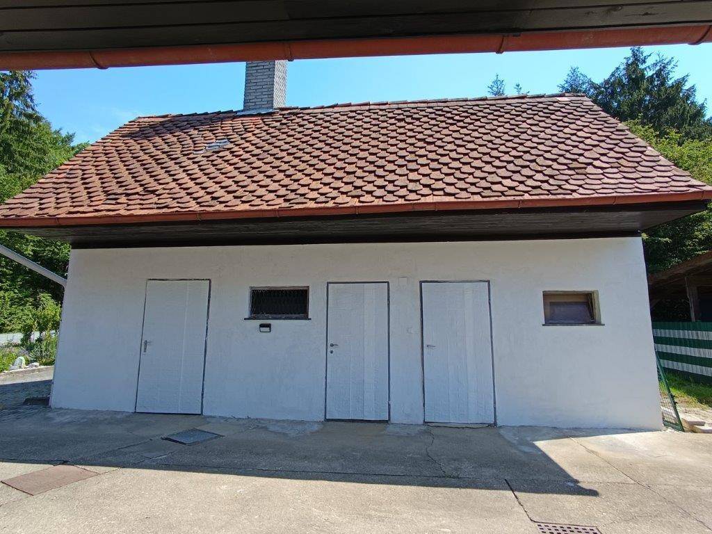 Kleines Nebengebäude mit mehreren Türen und einem Ziegeldach, umgeben von Natur.