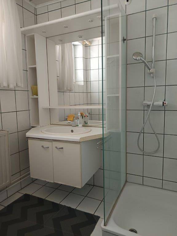 Badezimmer mit weißen Fliesen, Waschtischunterschrank, Spiegelschrank und Duschkabine.