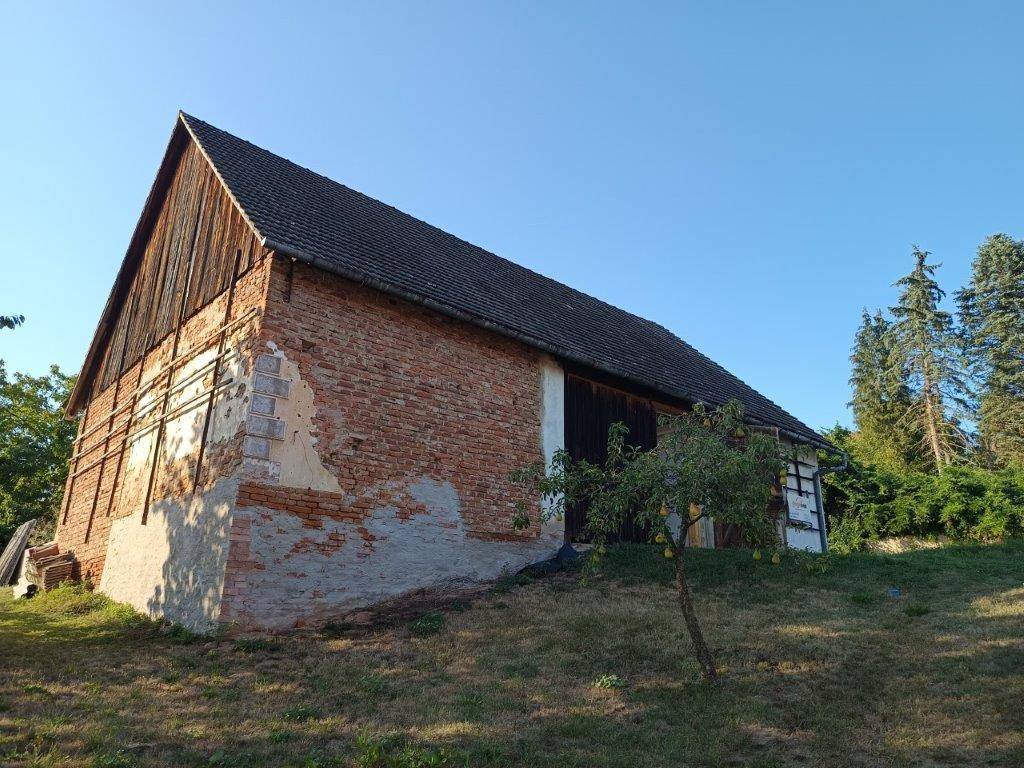 Lange Seite des alten Scheunengebäudes mit Ziegelmauerwerk, Holzbalken und einem kleinen Baum im Vordergrund.