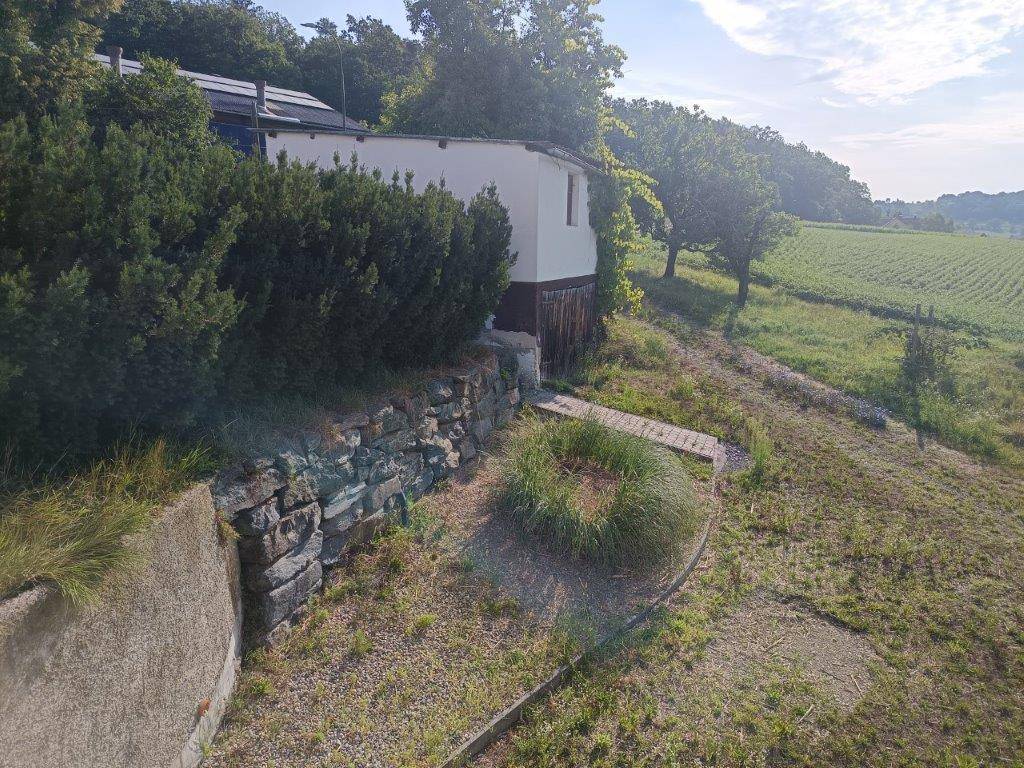 Gartenbereich mit Natursteinmauer und gepflegten Pflanzen, umgeben von grüner Landschaft.