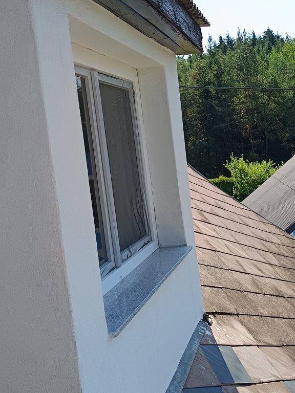 Fenster in einer weißen Fassade mit Dachziegeln und umgebenden Bäumen.