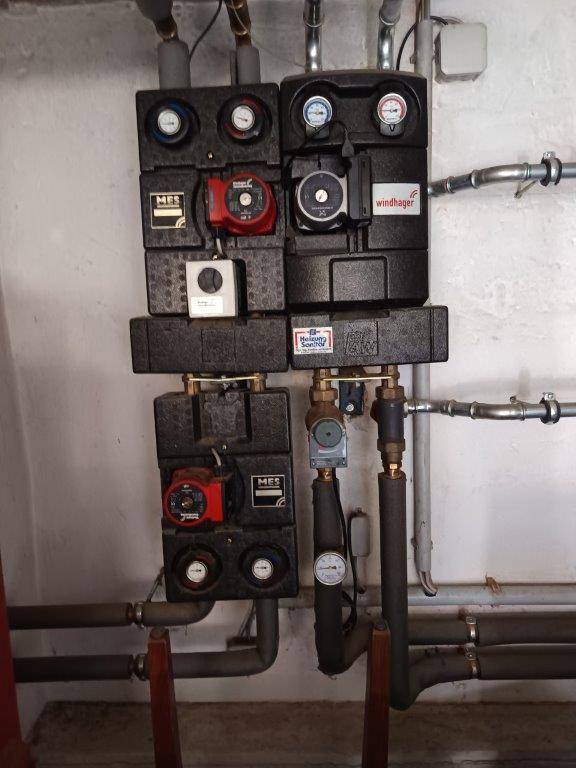 Heizungsverteiler mit Pumpen und Messgeräten an der Wand in einem Technikraum.