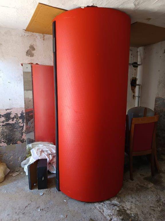 Großer roter Warmwasserspeicher in einem Kellerraum mit unfertigen Wänden.