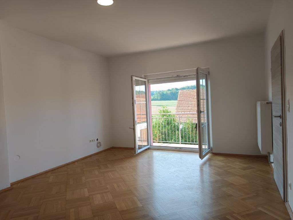 Helles Zimmer mit Parkettboden und französischem Balkon, bietet Blick auf Dächer und Natur.