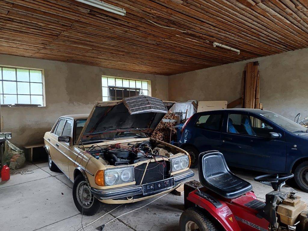 Innenansicht einer Garage mit einem älteren Mercedes mit offener Motorhaube und einem Rasenmäher.
