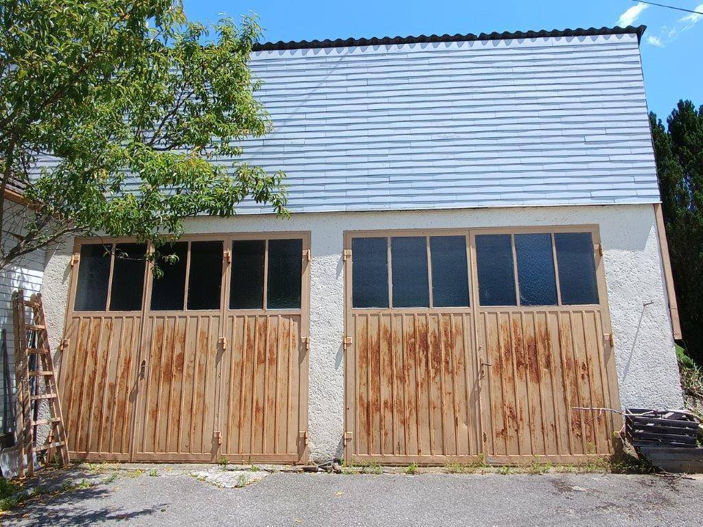Außenansicht einer Garage mit zwei großen, rostigen Holztoren und einem Wellblechdach unter blauem Himmel.