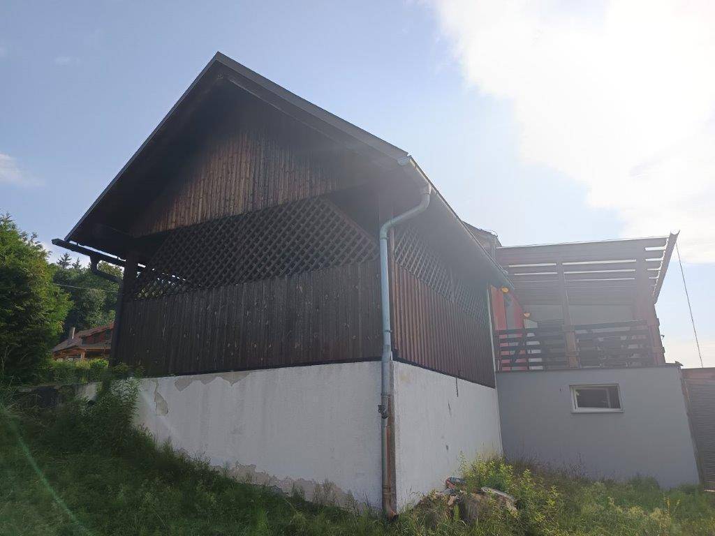 Seitenansicht eines Gebäudes mit Holzverkleidung und weißem Sockel, umgeben von Grün.
