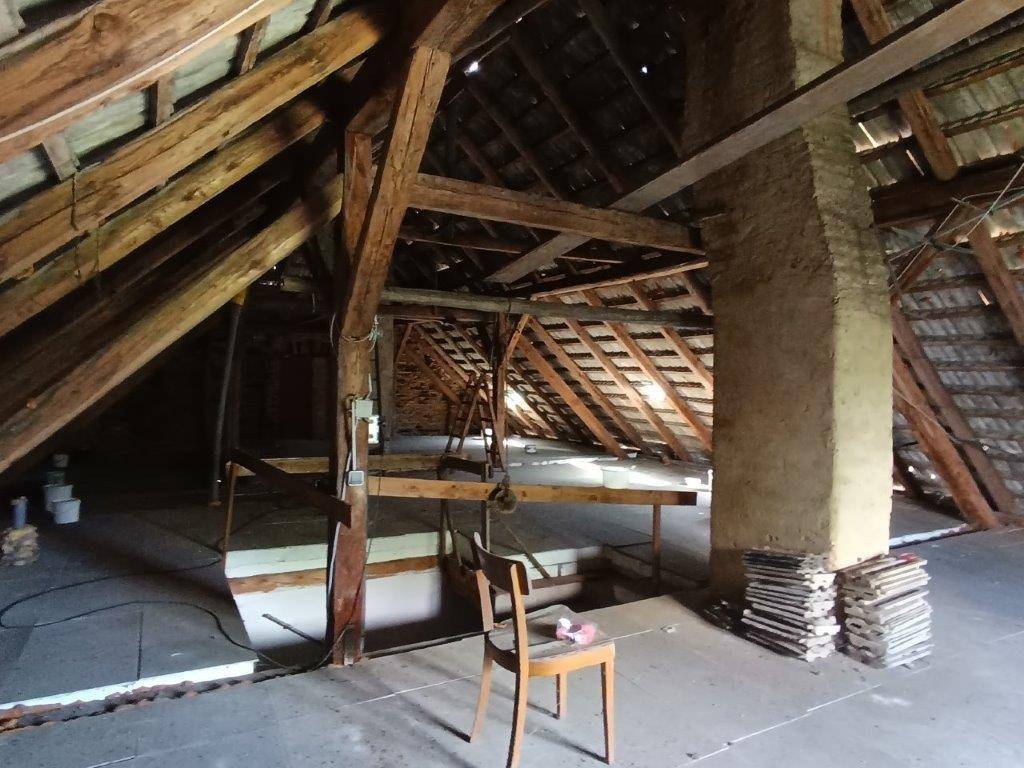 Unausgebauter Dachboden mit freiliegenden Holzbalken, einem gemauerten Kamin und gestapelten Baumaterialien.