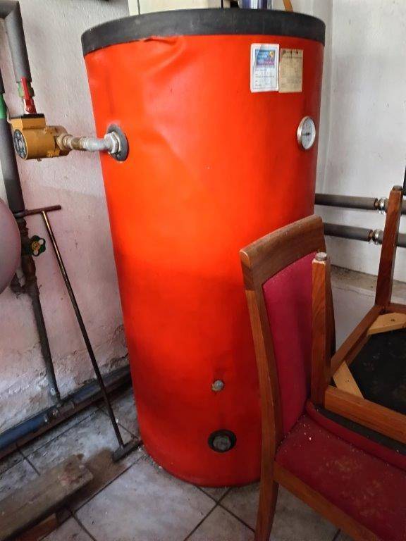 Roter Warmwasserspeicher mit Rohren und Messgeräten in einem Kellerraum.