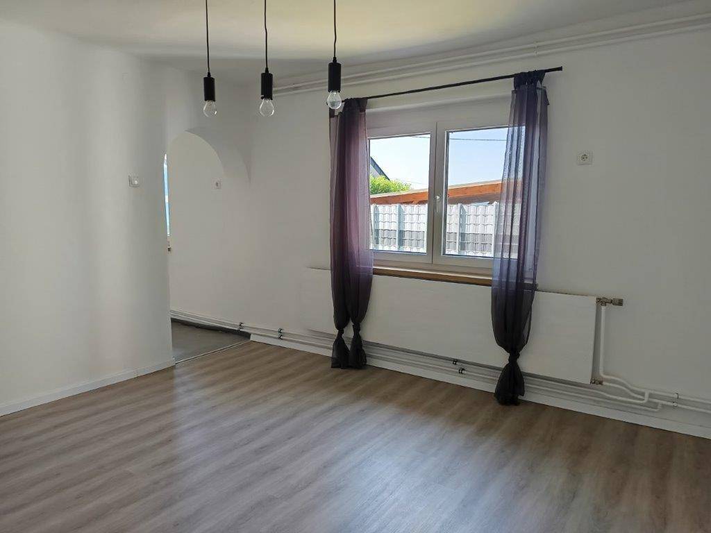 Heller Wohnbereich mit Laminatboden, weißen Wänden und einem Fenster mit Vorhängen.