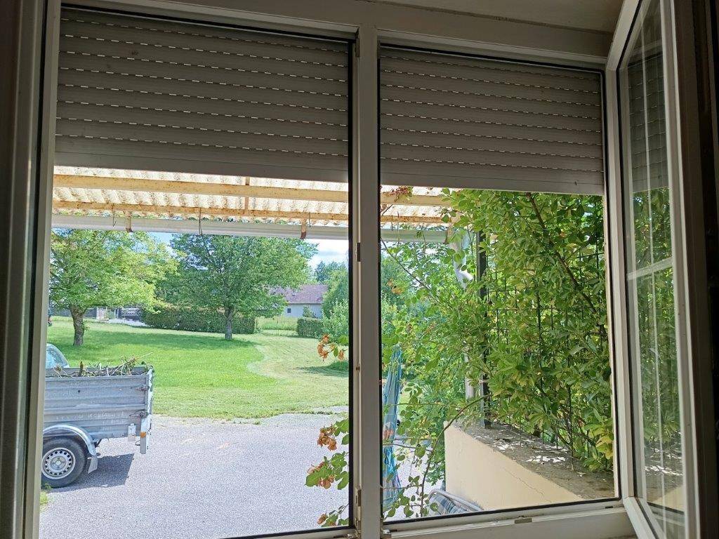 Fenster mit Blick auf einen Gartenbereich und einen Anhänger im Freien, mit Rollladen.