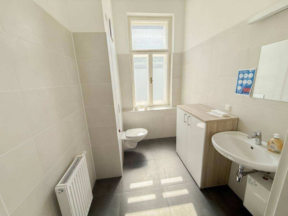 Modernes WC mit hellen Fliesen, wandhängendem WC, Einzelwaschbecken und Fenster.