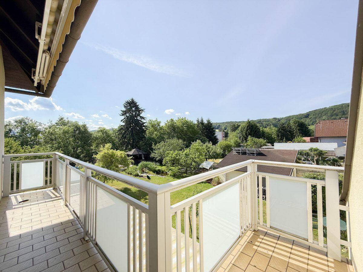 Geräumiger Balkon mit Fliesenboden, weißem Geländer und weitem Blick ins Grüne.