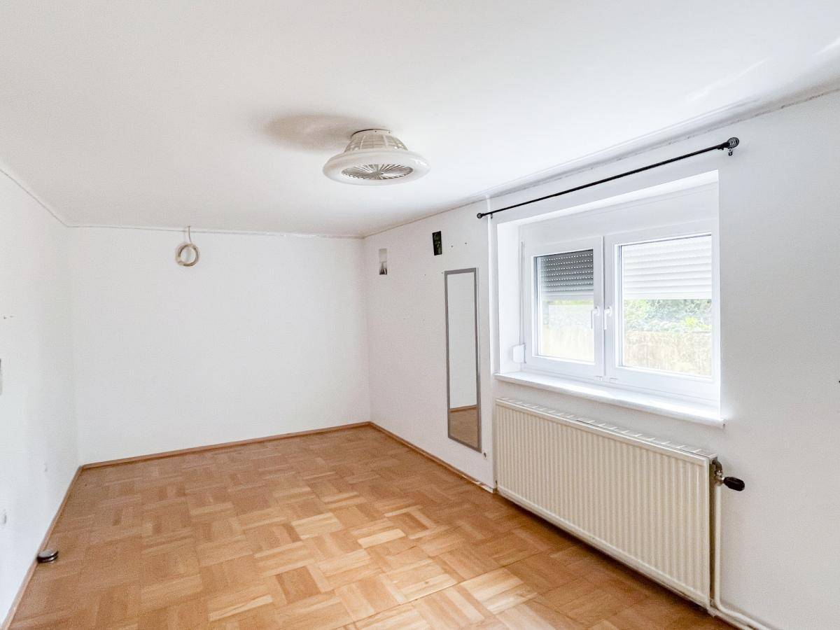 Leerer Raum mit hellem Parkettboden, großem Fenster und Heizkörper.