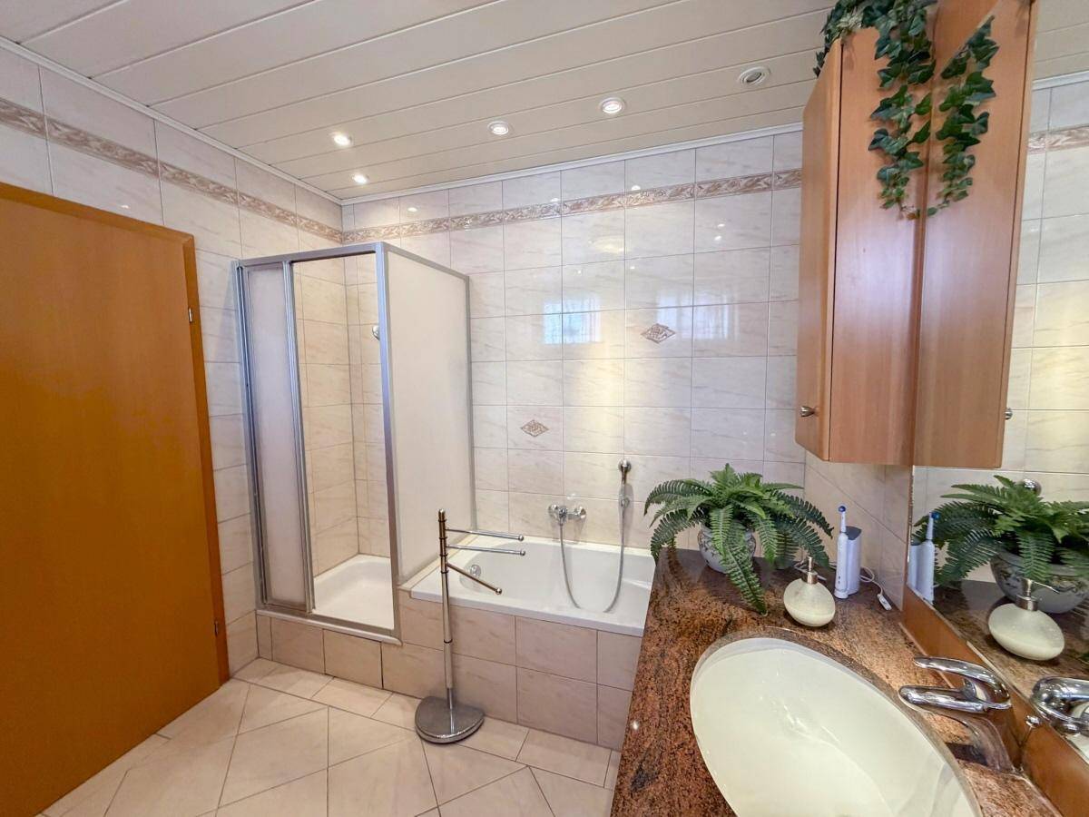 Badezimmer mit Badewanne, Duschkabine und Waschtisch, umgeben von hellen Fliesen und Holzmöbeln.