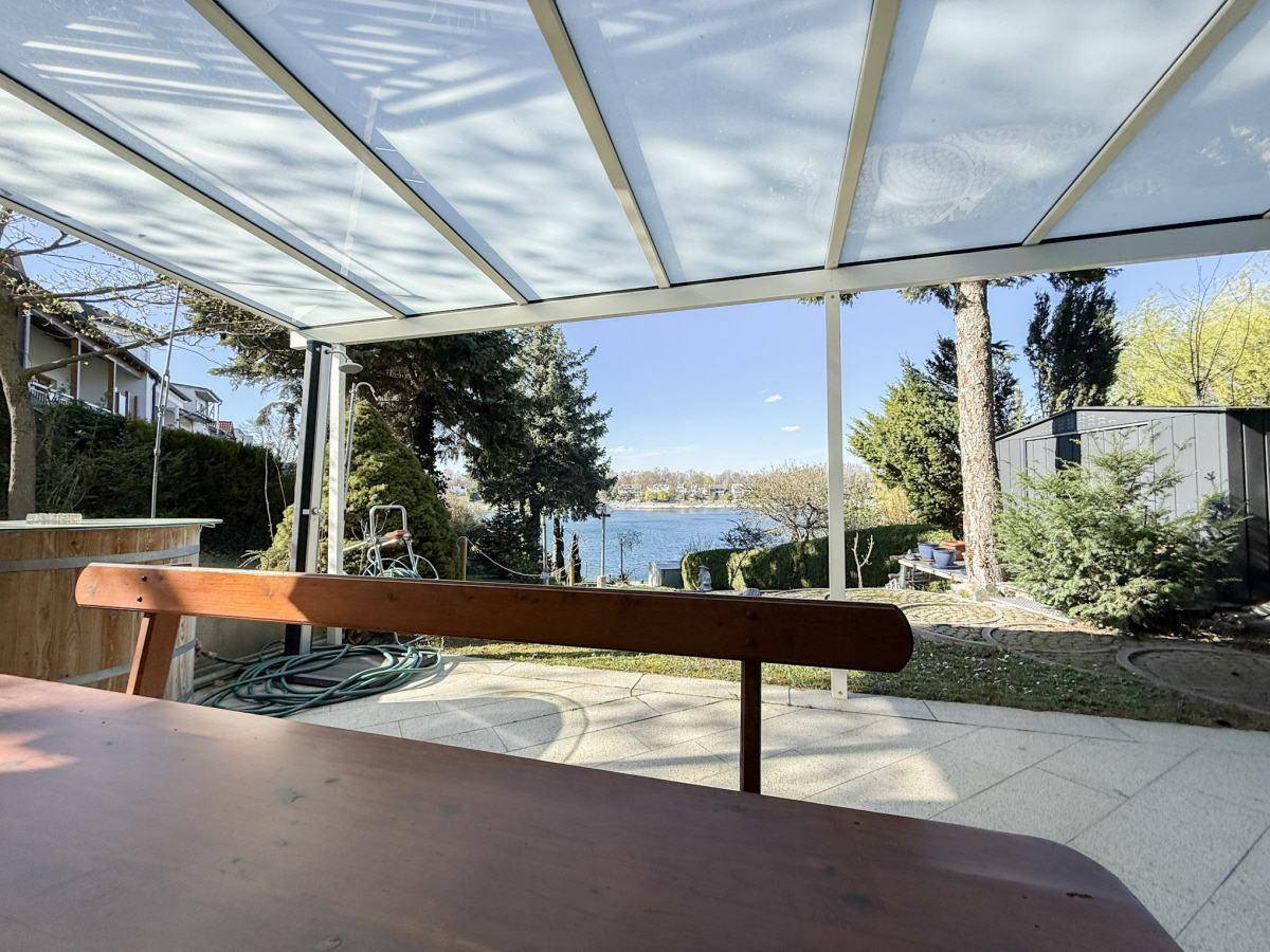 Überdachte Terrasse mit Holztisch und Bänken, die einen malerischen Blick auf den See bietet.