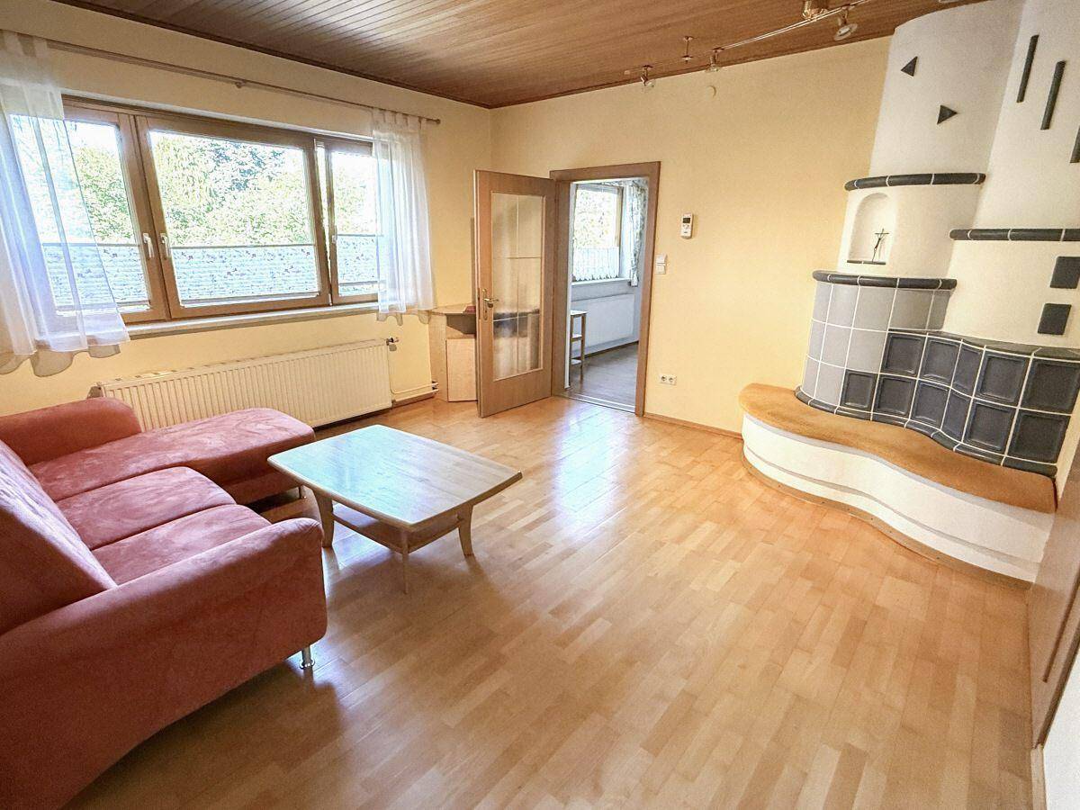 Gemütliches Wohnzimmer mit traditionellem Kachelofen, hellem Parkettboden und Zugang zu weiteren Räumen.