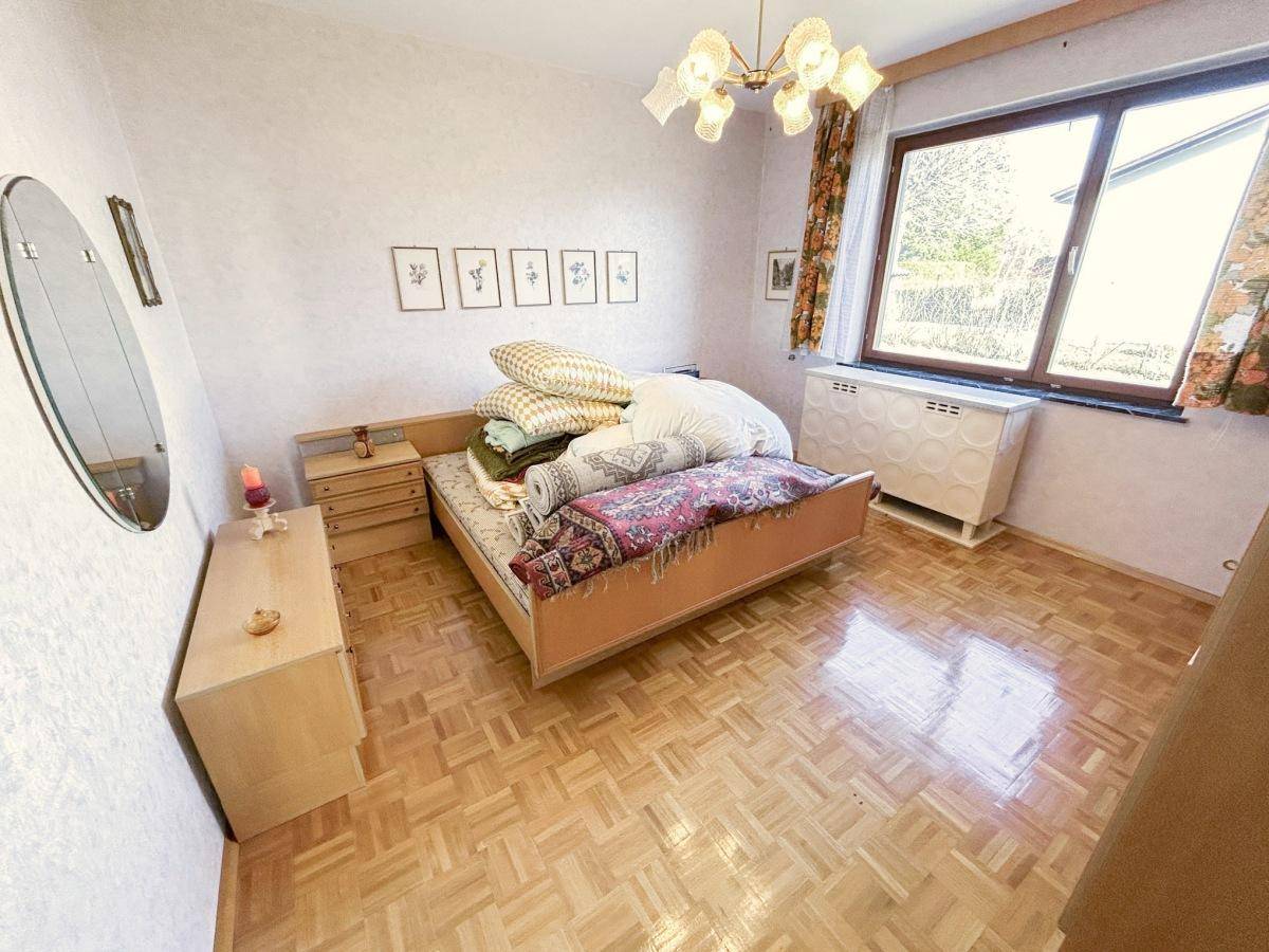 Schlafzimmer mit Parkettboden, großem Fenster und einfacher Einrichtung.