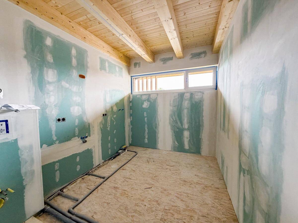 Rohbau eines Badezimmers mit Holzbalkendecke, horizontalen Fenstern und vorbereiteten Anschlüssen.