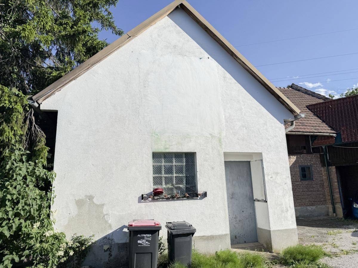 Weiß verputztes Nebengebäude mit Giebeldach, einem kleinen Fenster und Mülltonnen im Vordergrund.