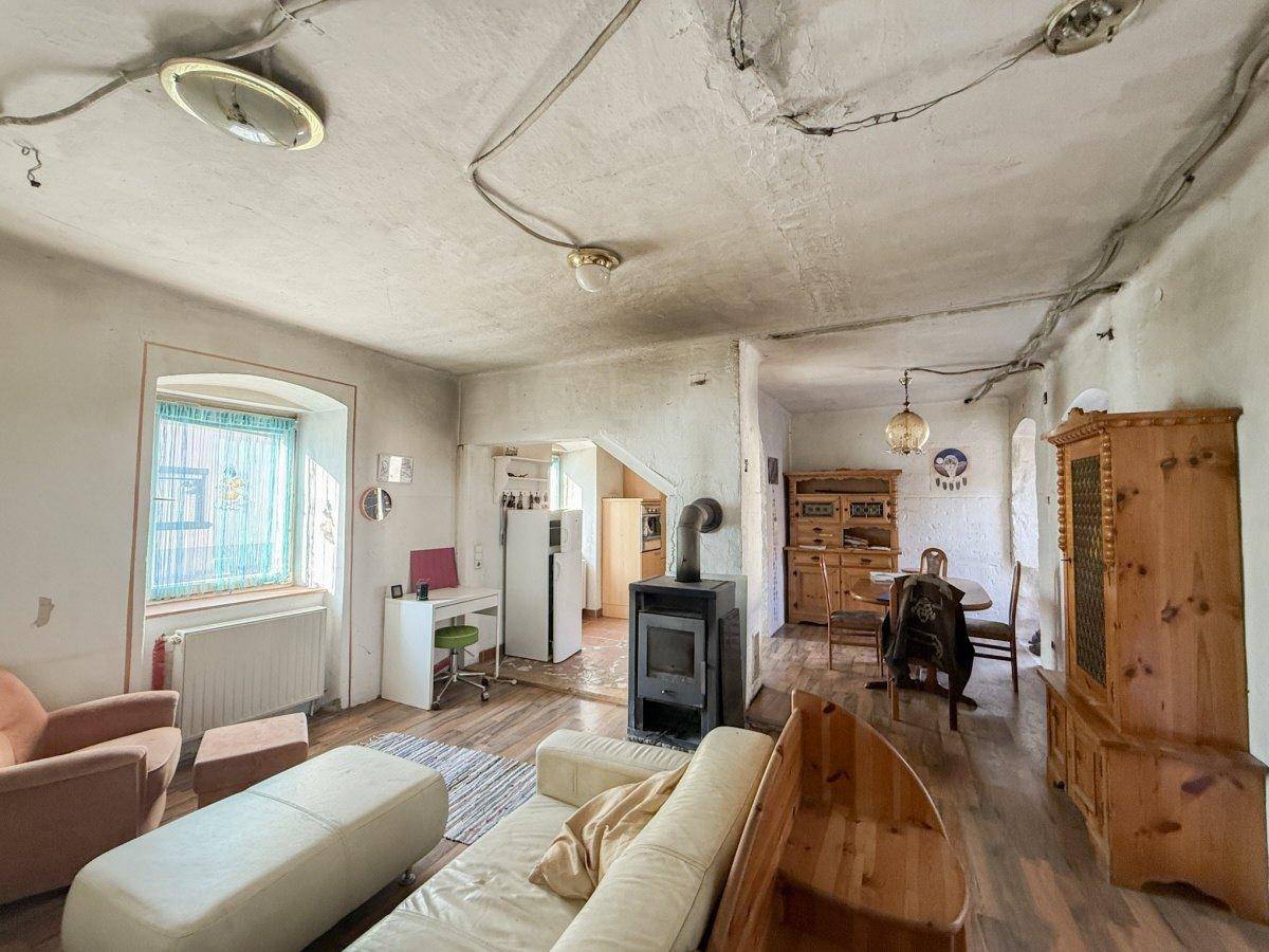 Kombinierter Wohn- und Essbereich mit Sofa, Schreibtisch, Holzofen und Esstisch, mit sichtbaren Elektroleitungen.