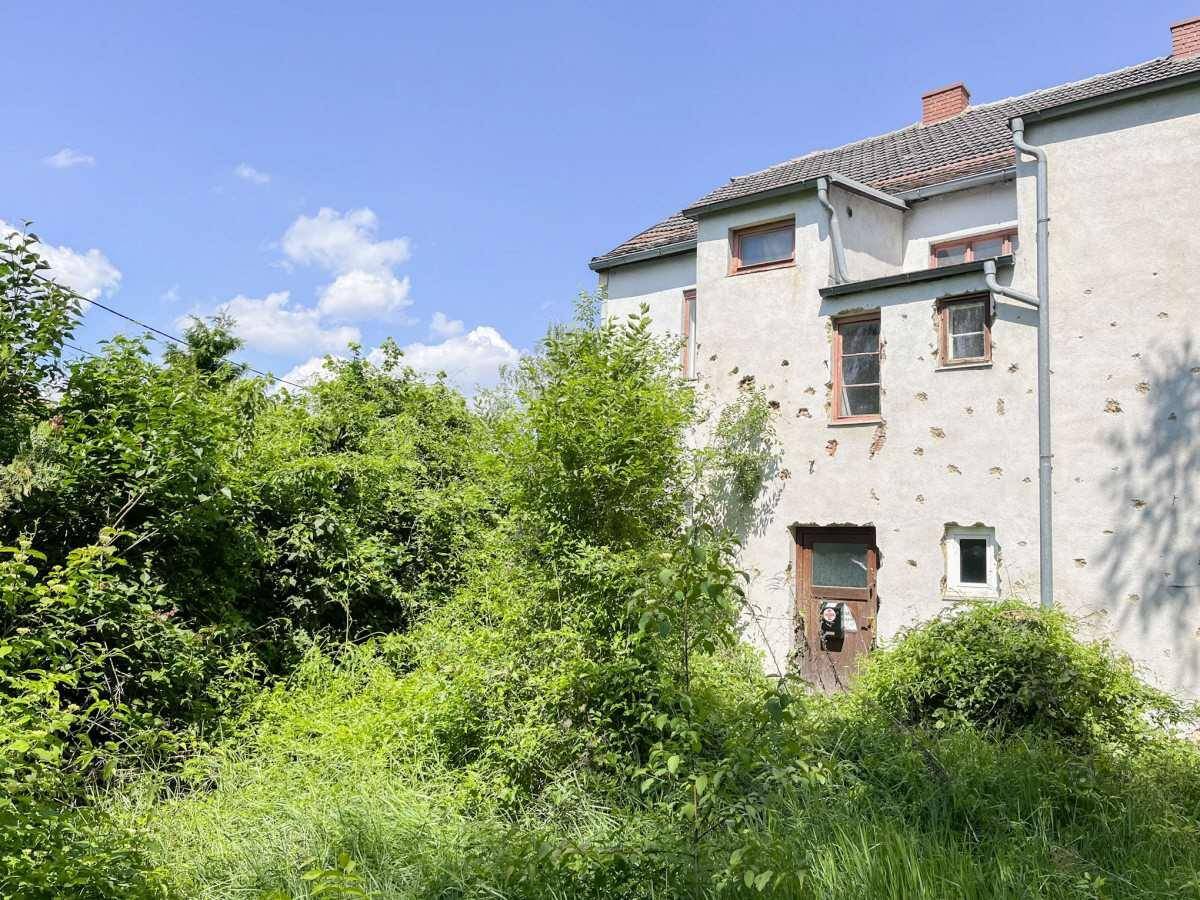 Vorderansicht des Hauses mit beschädigter Fassade und überwuchertem Vorgarten bei Sonnenschein.