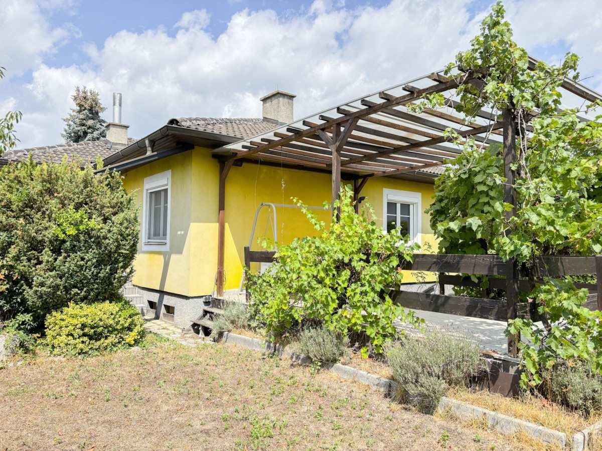 Gelbes Einfamilienhaus mit überdachter Terrasse und Gartenbereich unter blauem Himmel.