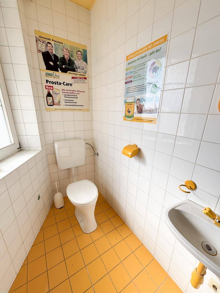 Kleine Toilette mit gelben Bodenfliesen und weißen Wandfliesen, ausgestattet mit einem Fenster.
