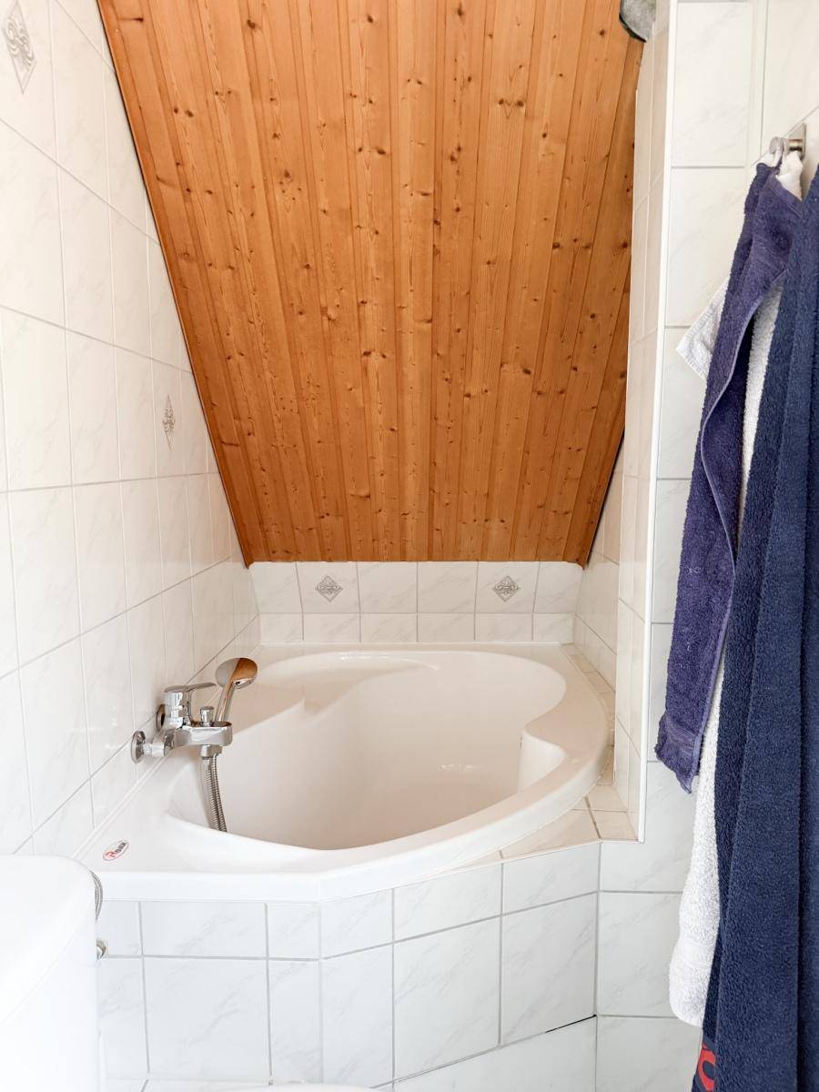 Badezimmer mit Eckbadewanne, weißen Fliesen und einer charmanten Holzdecke im Dachgeschoss.