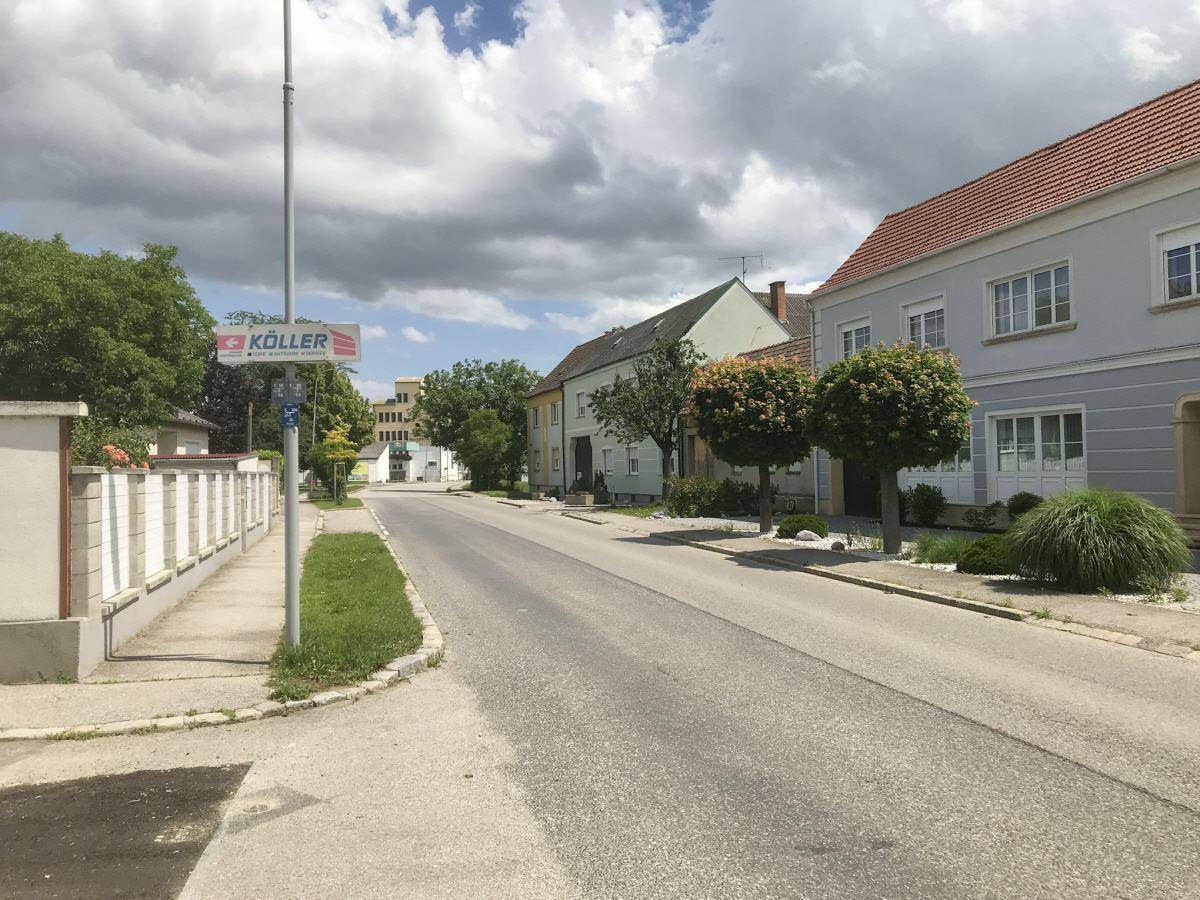 Dorfstraße mit gepflegten Häusern und einem Werbeschild unter bewölktem Himmel.