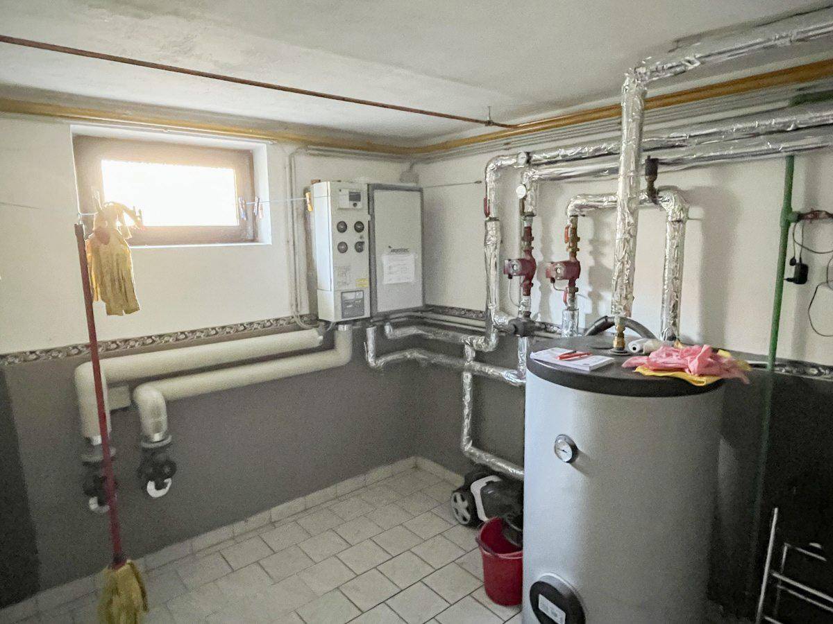 Heizungsraum im Keller mit Warmwasserspeicher, Rohren und einem kleinen Fenster.