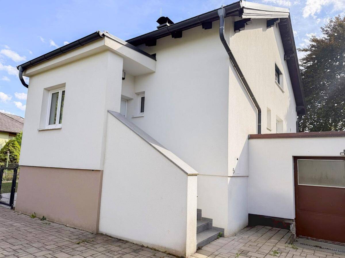 Ansicht des Hauses von außen mit weißer Fassade, Satteldach und Zugang über eine Treppe.