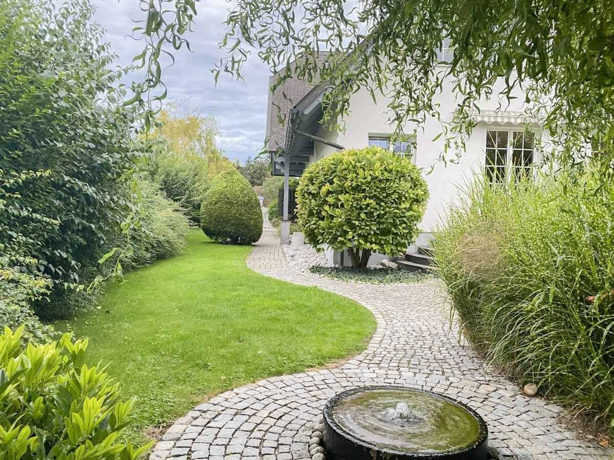 Geschwungener Gartenweg mit Brunnen und Blick auf das Haus, umgeben von grünen Büschen.