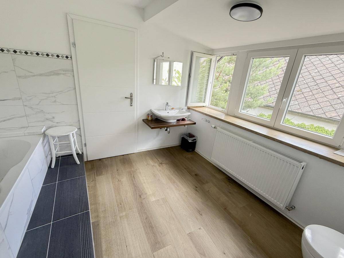 Helles Badezimmer mit Waschbecken, Badewanne und großen Fenstern mit Blick auf die Dächer.