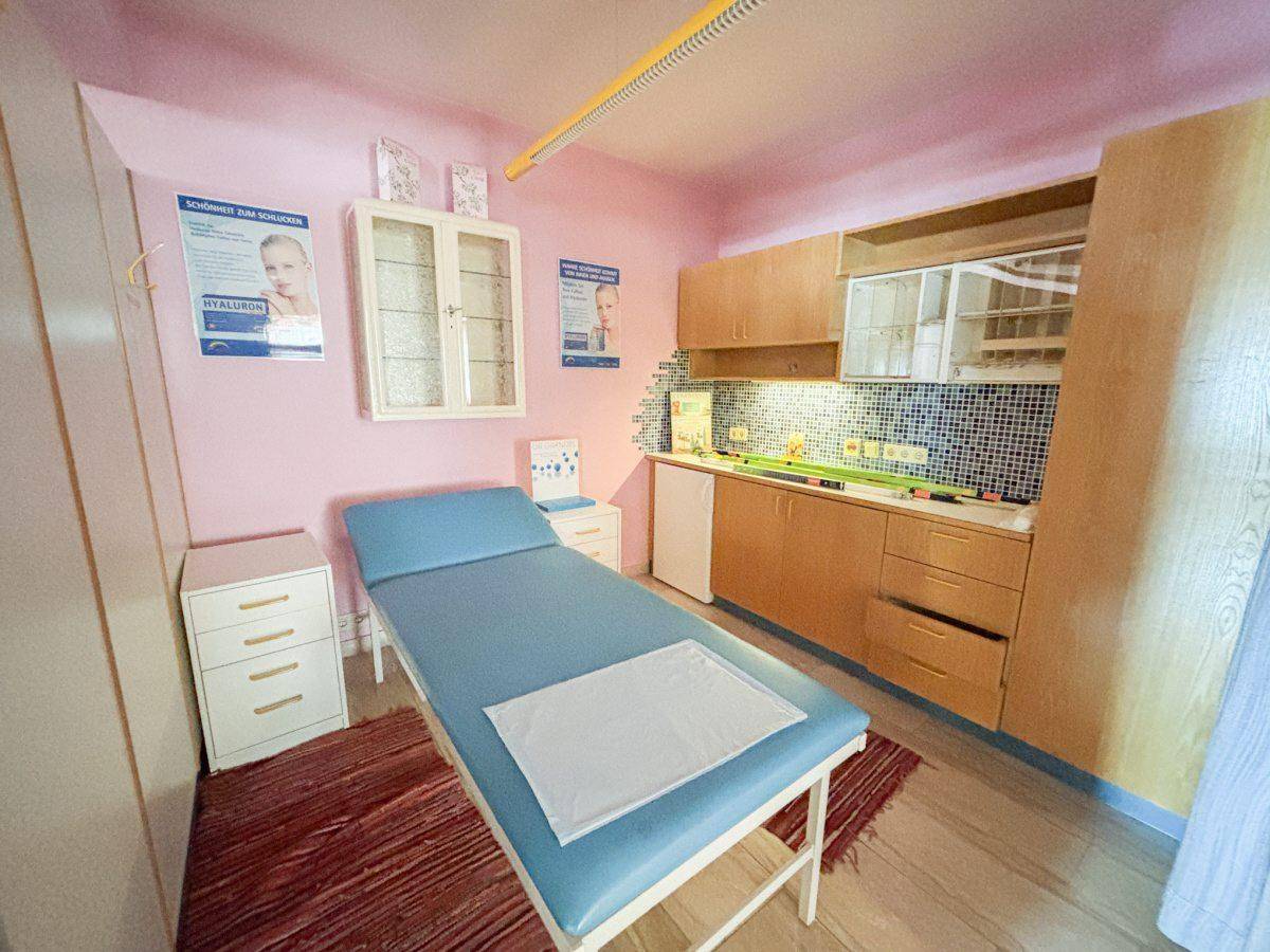 Kleine Küchenzeile mit Spüle, Kühlschrank und Holzschränken, integriert in einen Behandlungsraum.