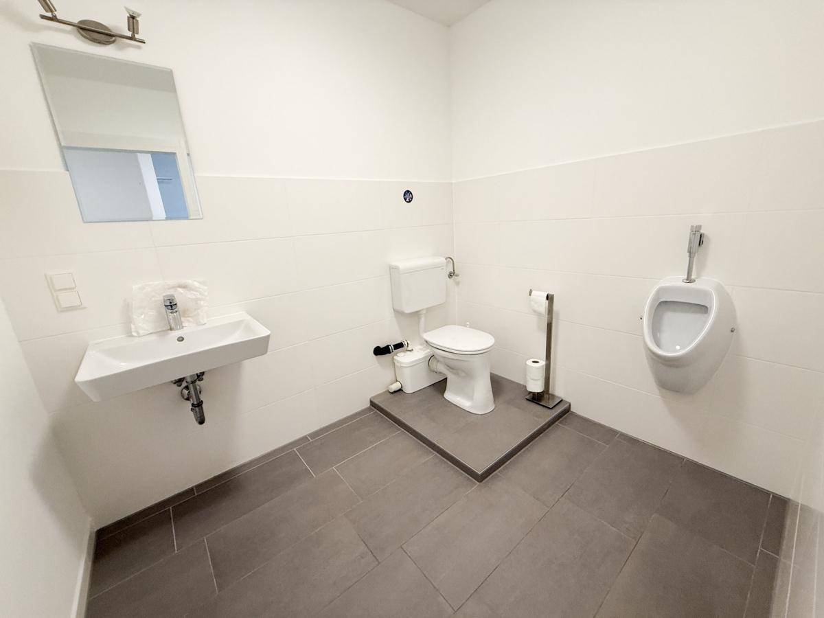 Geräumiges WC mit Waschbecken, Toilette und Urinal, ausgestattet mit grauen Bodenfliesen.